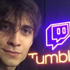 Il volto di un giovane uomo con capelli scuri e occhi castani, accanto a un'insegna al neon di Twitch e "Tumblu". Chissà chi è Blur?