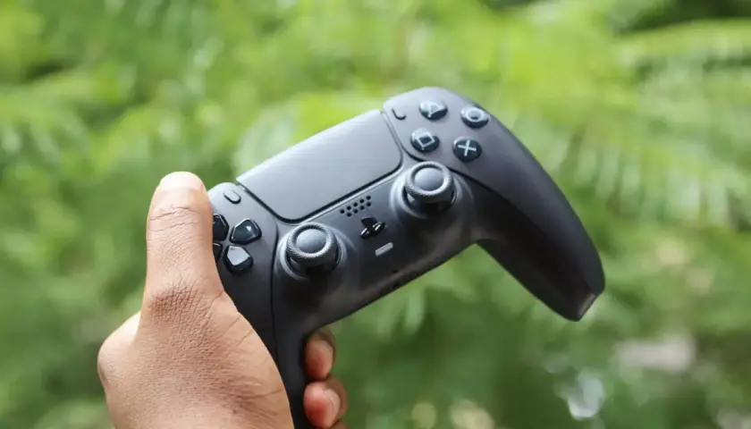 Mano che impugna il tasto crea PS5, pronto per nuove avventure videoludiche tra il verde della natura.
