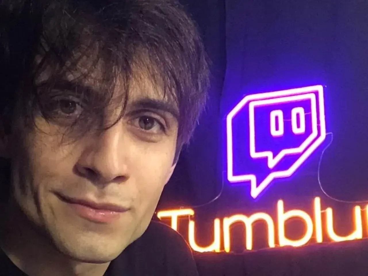 Il volto di un giovane uomo con capelli scuri e occhi castani, accanto a un'insegna al neon di Twitch e "Tumblu". Chissà chi è Blur?